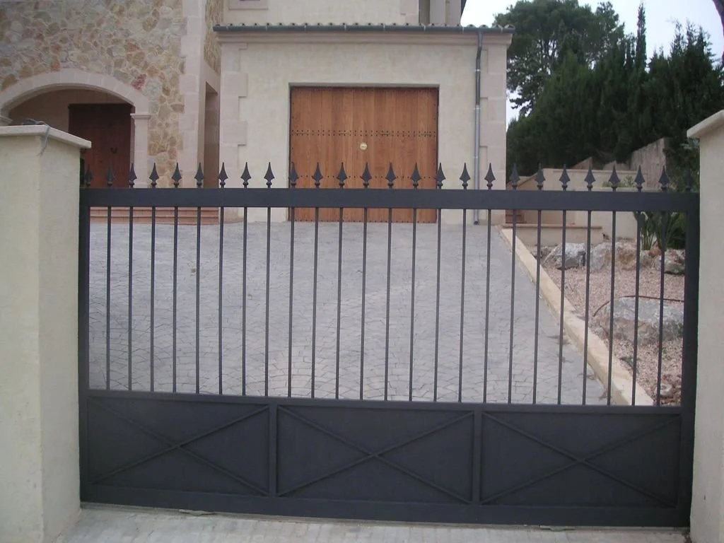 Una puerta metálica negra con rejas que cierra una casa de piedra con garaje de madera y un camino de piedra en una casona rodeada de árboles.