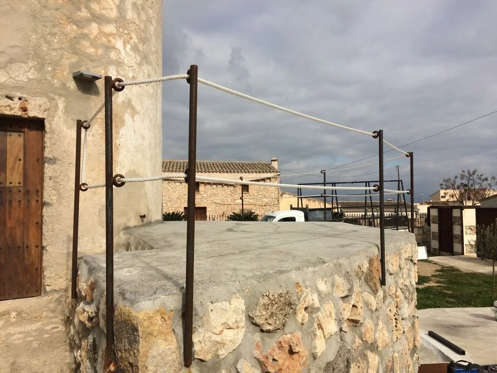 Estructura de cuerda para escalar en un poste de piedra, en un patio con casas antiguas y cielo nublado.