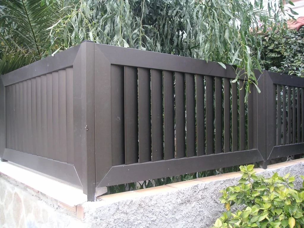 Barandilla negra de metal en un muro de piedra, con plantas verdes alrededor.