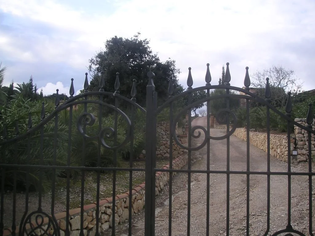 Una puerta negra de rejas con picos puntiagudos, que da a un camino de grava rodeado de muros de piedra y vegetación, con árboles y cielo nublado en el fondo.