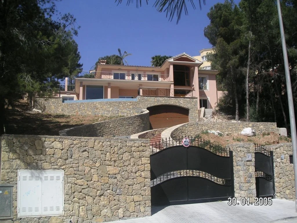 Casa grande de varios pisos en una colina, con puerta negra de hierro, muros de piedra y área ajardinada, rodeada de árboles y un cielo despejado.