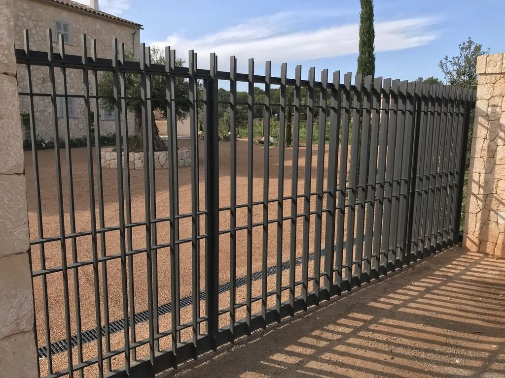 Valla de metal negro con sombra en el suelo, en un área exterior con tierra y plantas, cerca de una estructura de piedra.