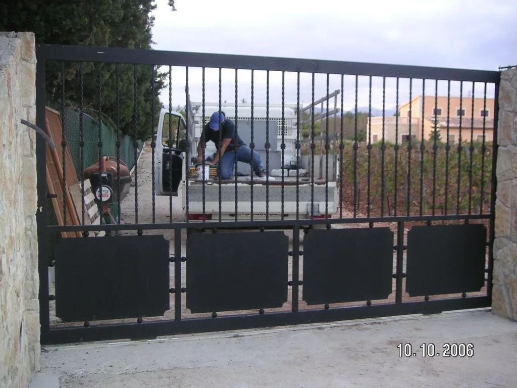 Persona trabajando en la parte trasera de un vehículo en un área cercada con muro de piedra y reja de metal, con herramientas y objetos almacenados al lado.