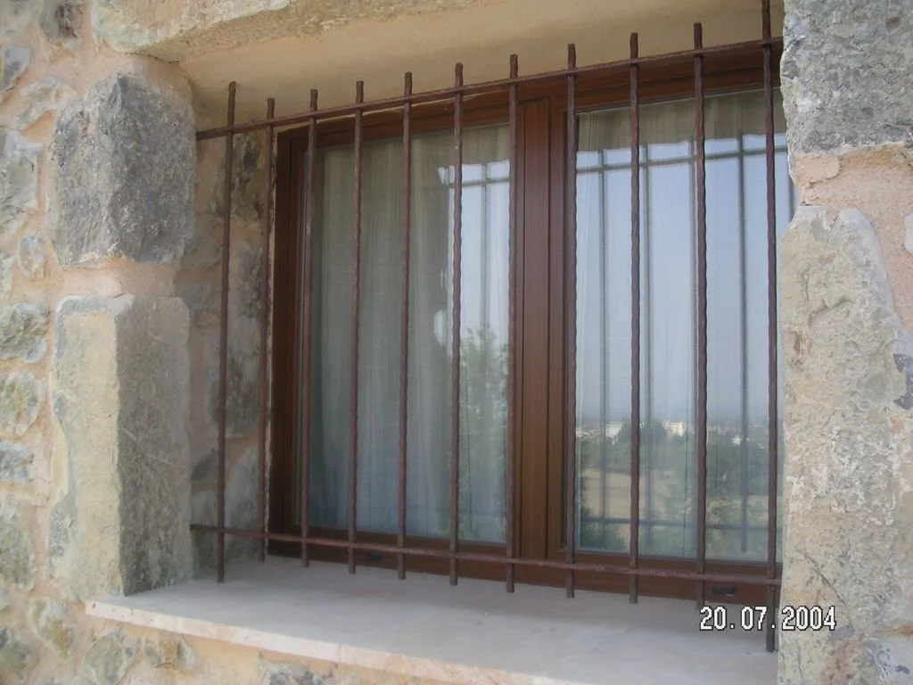Ventana de madera con reja de hierro en una pared de piedra, con vista a un paisaje exterior y cortinas blancas adentro, fecha 20-07-2004.