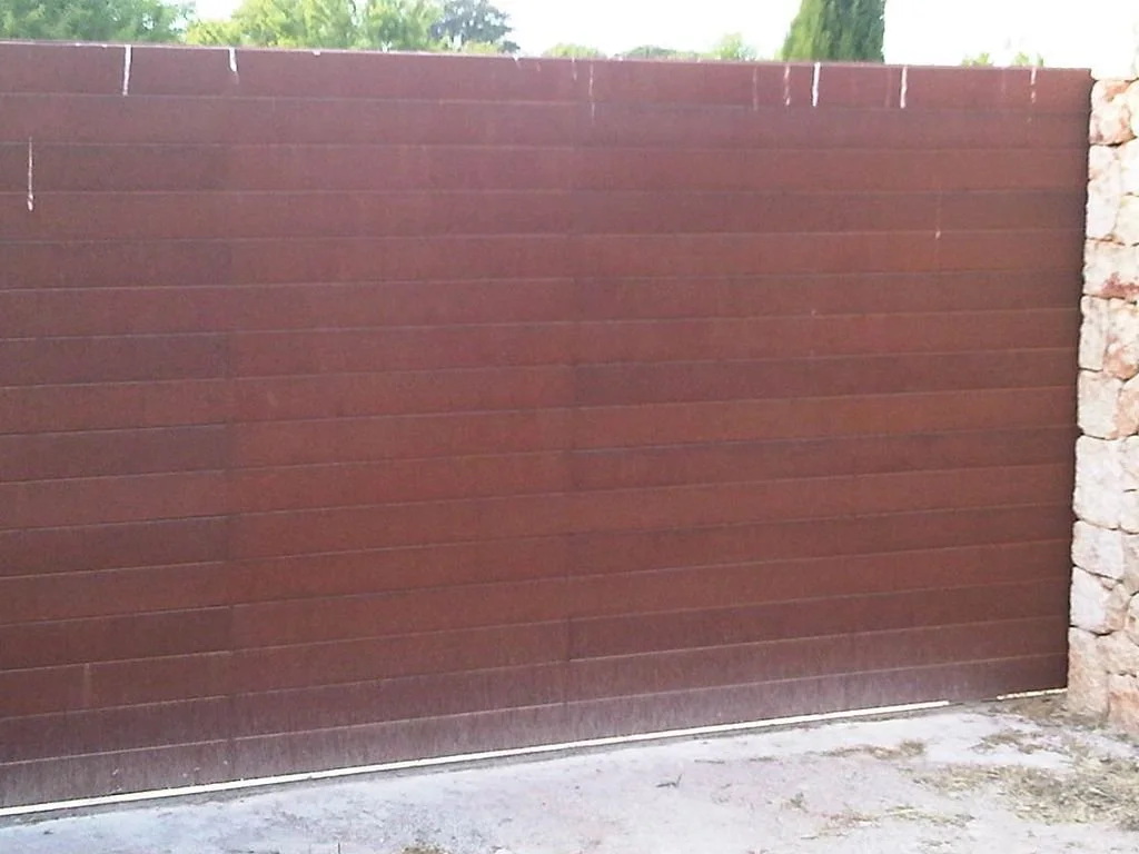 Portón de hierro marrón con muro de piedras a la derecha y árboles verdes detrás.