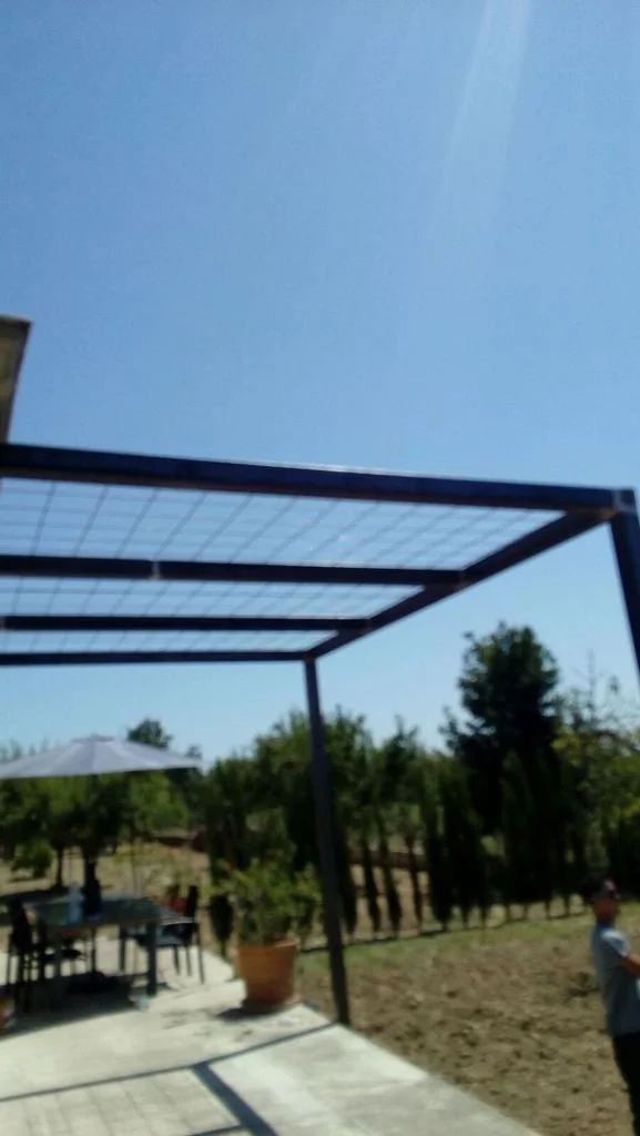 Área exterior con una estructura de pérgola sin paredes, un comedor con sombrilla y sillas, y árboles en el fondo.