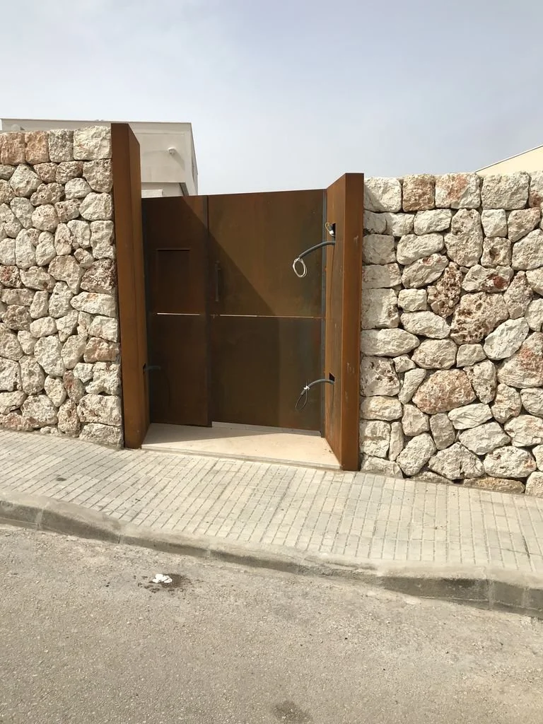 Puerta de metal café sin candado en muro de piedra con acera y calle en primer plano.