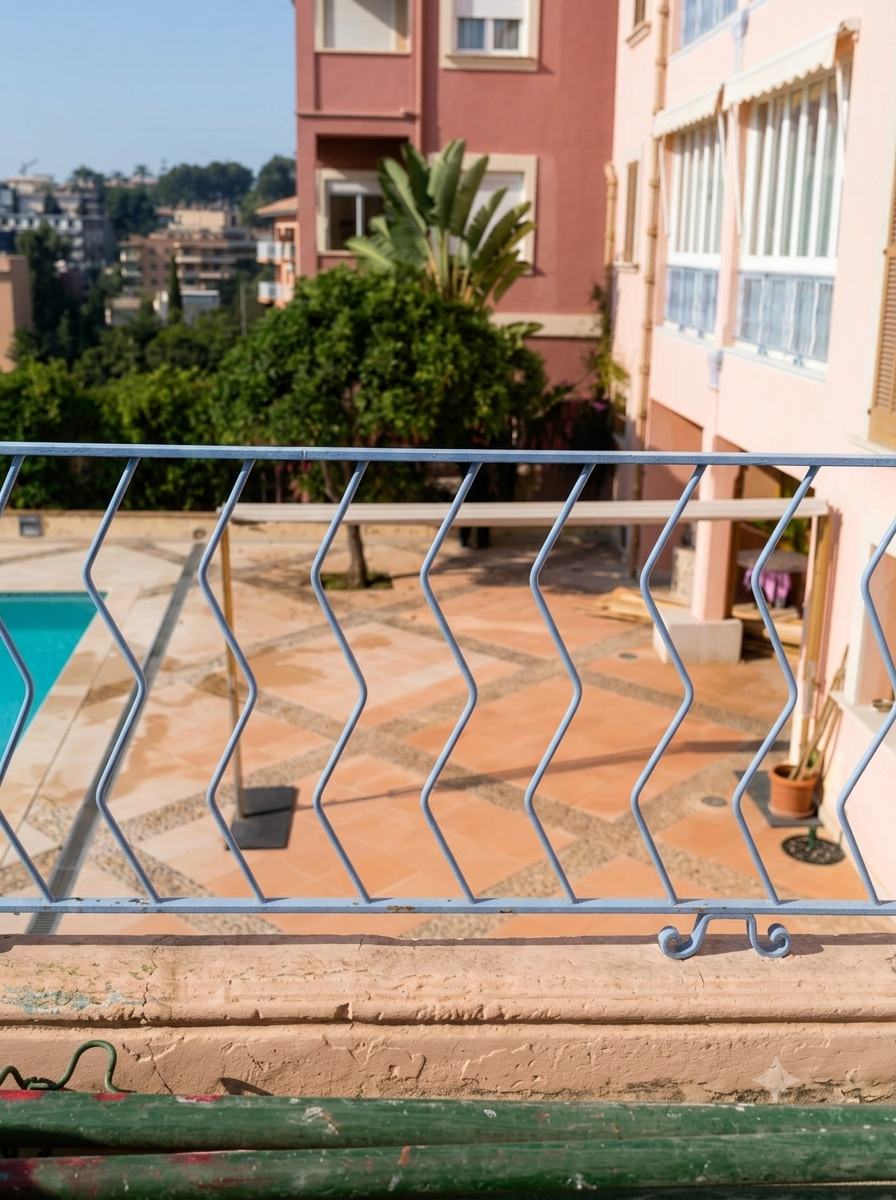 Vista desde un balcón con barandilla de metal, piso de terrasse con baldosas, piscina y plantas, incluyendo una palmera, en un área residencial con edificios de colores pastel.