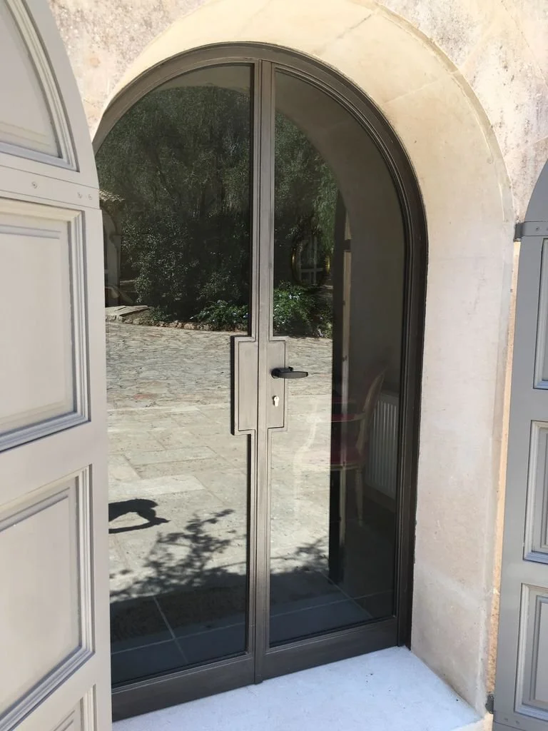 Puerta de vidrio con marco gris y arco en una pared de piedra beige.