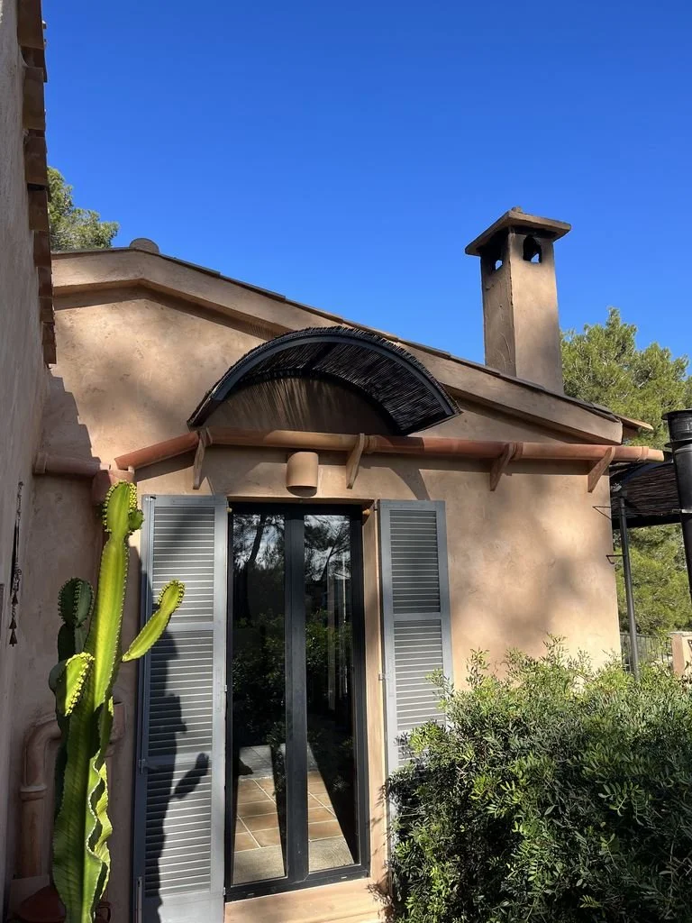 Casa con fachada de color beige, ventanas con contraventanas grises, azulejo en el suelo, cactus junto a la puerta de vidrio, chimenea en el techo y un cielo azul despejado.