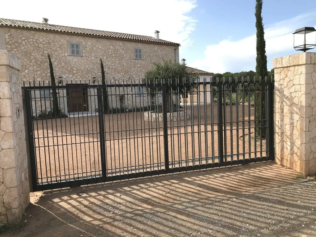 Puerta de reja negra en entrada de casa de piedra con muro de piedra a cada lado y caminos de tierra