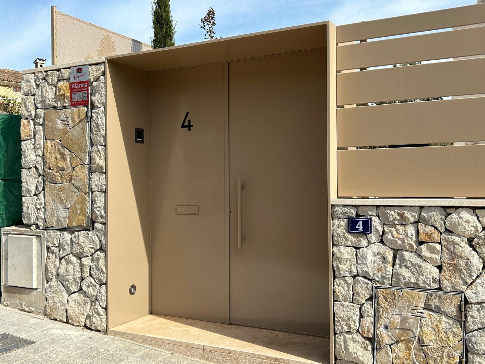 Entrada de una casa con muro de piedra y una puerta metálica beige con el número 4, señalización de alarma y buzón.