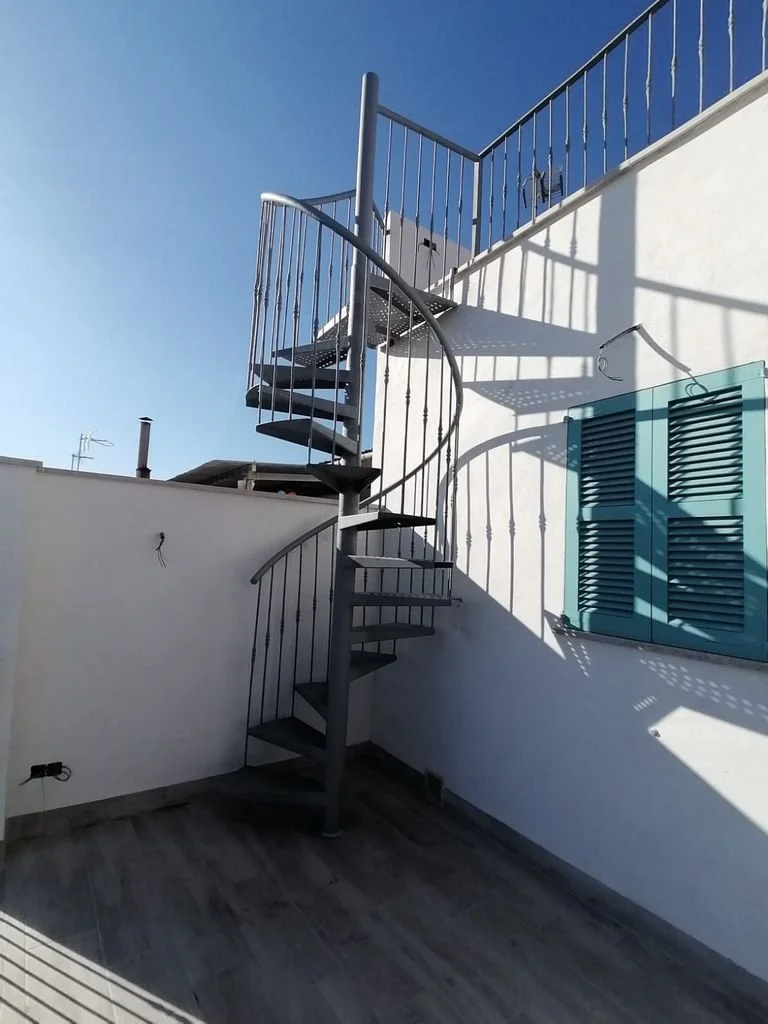 Escalera de metal en un patio con paredes blancas y puerta de persiana azul, con sombra proyectada por la barandilla y la estructura en un día soleado.