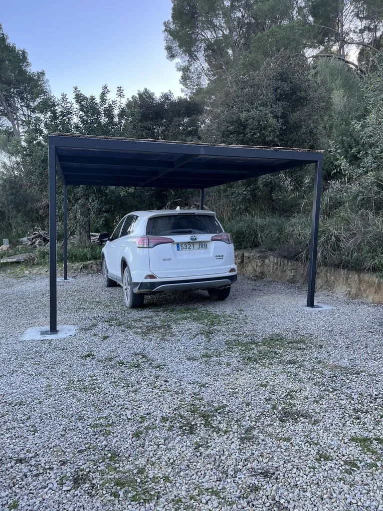 Cochera sin espacio para un vehículo, con techo metálico, en un área de grava, y un coche estacionado parcialmente en ella.