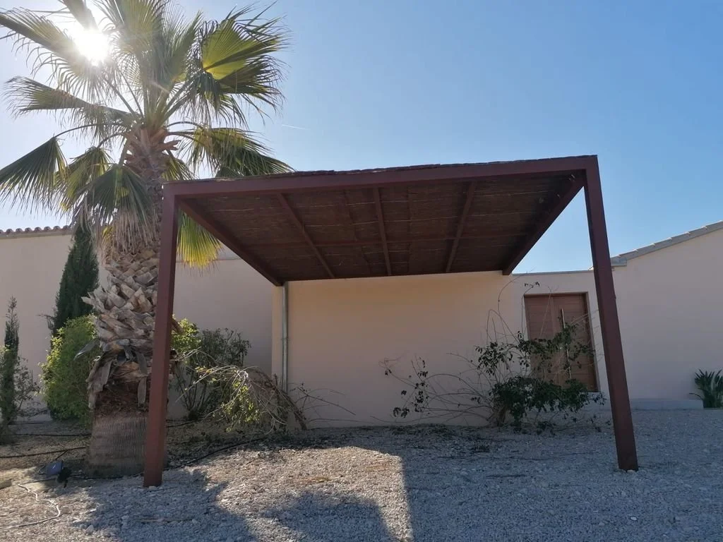 Estructura de sombra con techado de madera, rodeada de plantas y palmeras, en un área exterior con suelo cubierto de piedras.