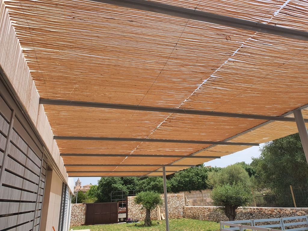 Techo de pérgola con sombra de madera y columnas metálicas en un jardín con árboles y paredes de piedra.