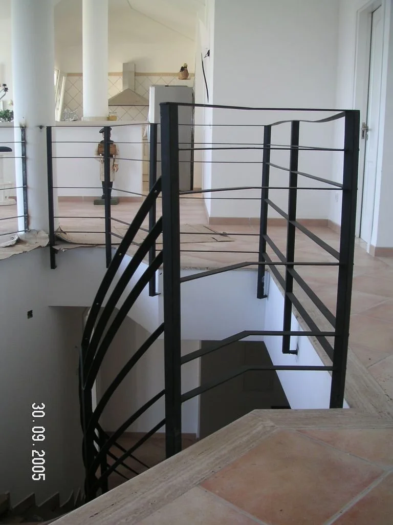 Un espacio interior en construcción con barandales de metal negro alrededor de una escalera que desciende a un nivel inferior. En el fondo, una cocina con extractor y una nevera. El suelo es de baldosas de color beige y hay una puerta de vidrio y ven