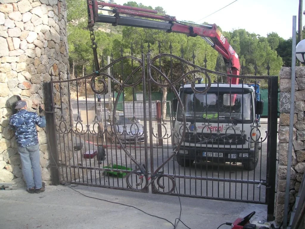 Un trabajador instalando un portón de hierro con diseño decorativo en una entrada de reja, con un camión de grúa desde adelante ayudando en la instalación.
