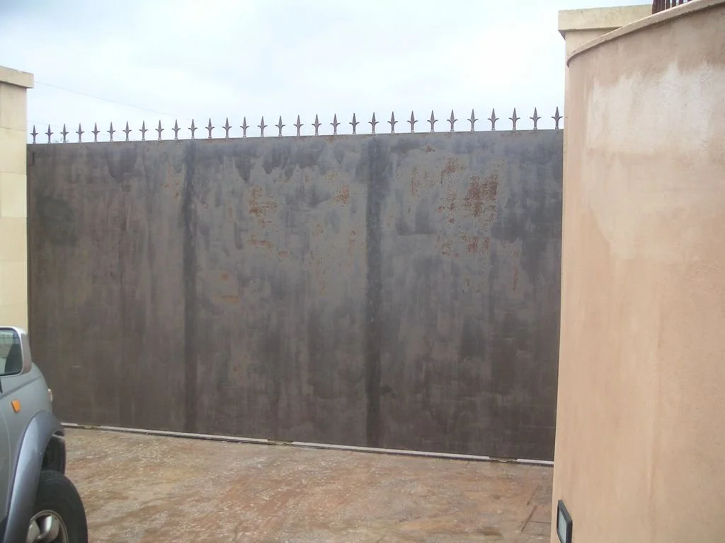 Muro de metal gris oscuro con picos en la parte superior, delgado y alto, en un entorno exterior, con una pared de color beige a la derecha y parte de un vehículo en la esquina inferior izquierda.