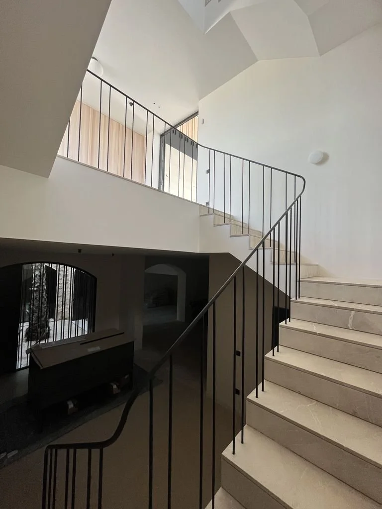 Escalera de mármol y barandilla de metal en un interior moderno de casa con paredes blancas y techos altos.