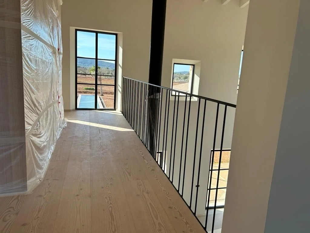 Interior de una casa en construcción con ventanas y barandal de metal, piso de madera sin terminar y un área con plástico protector.