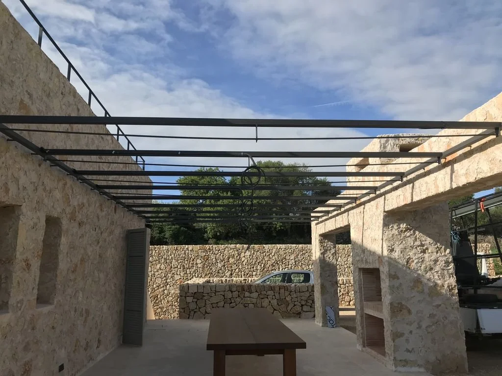 Estructura en construcción en un patio con paredes de piedra y una mesa de madera al centro, con un coche estacionado al fondo y un cielo con nubes.