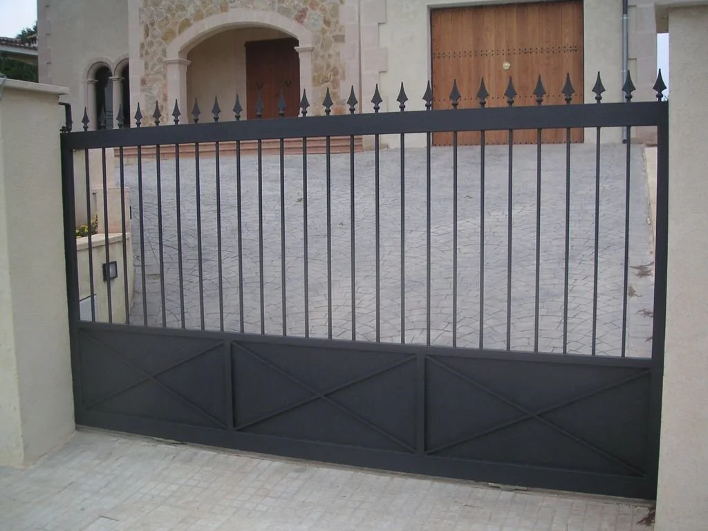 Reja negra de metal en la entrada de una casa con muro de piedra y puertas de madera al fondo.