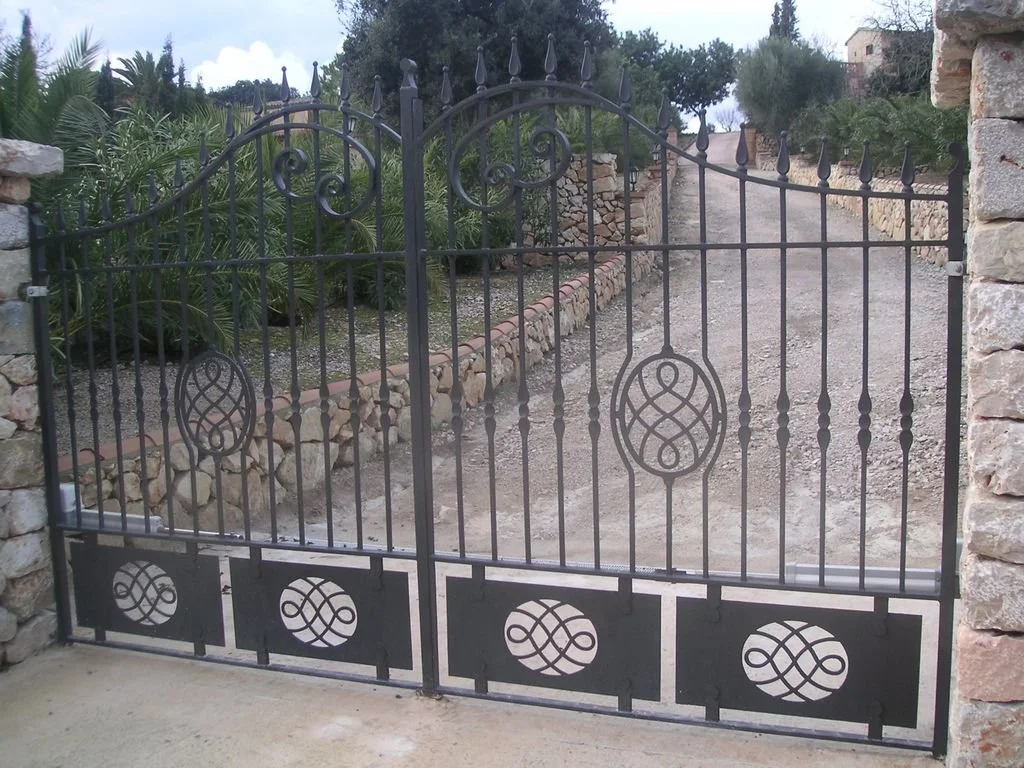 Puerta negra de rejas de metal con diseño decorativo, ubicada entre muros de piedra, con un camino de tierra y vegetación en el fondo.