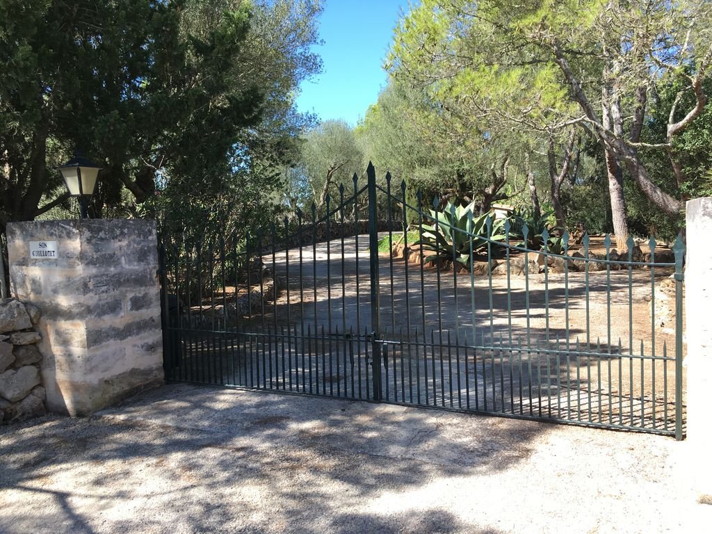 Puerta de hierro negra que da acceso a un camino rodeado de árboles y plantas en un entorno natural, con muros de piedra a los lados y sombra en el suelo.