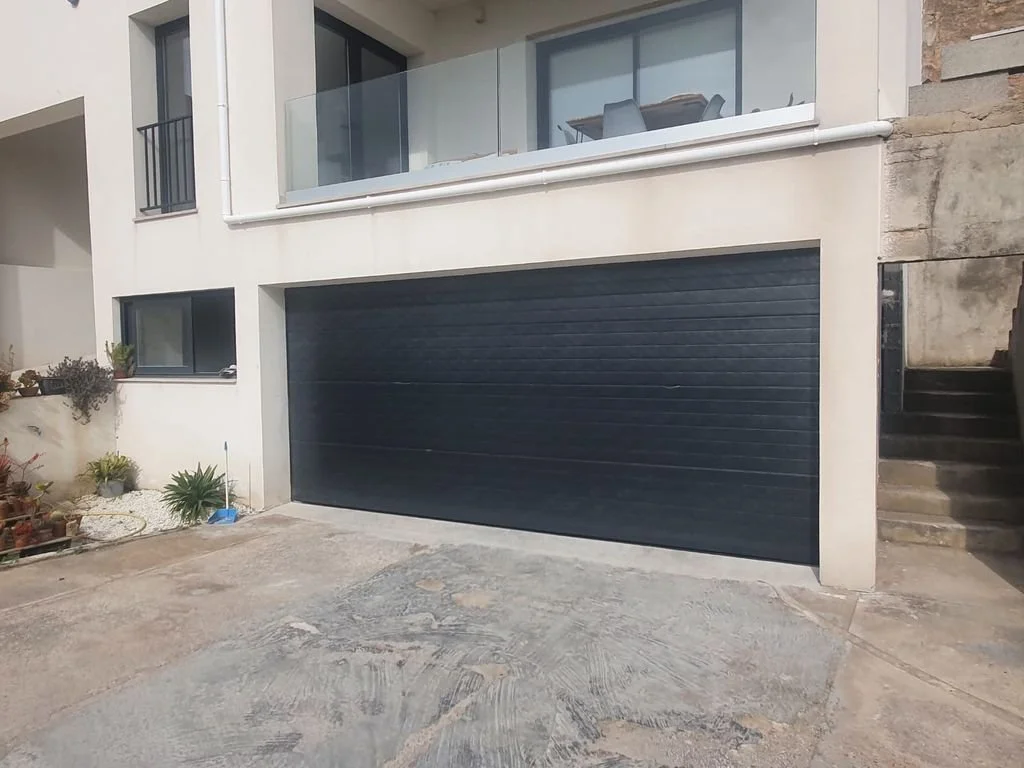 Fachada de una casa moderna con cochera de portón negro y ventanas grandes con balcón de vidrio en segundo piso, con plantas en el costado izquierdo y escaleras a la derecha.