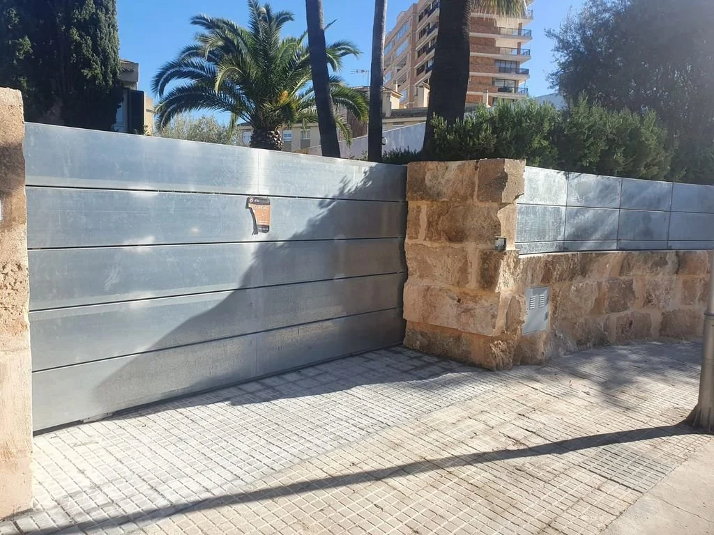 Cerca de muro con estructura de metal y piedra en una calle con arboles y edificios altos en el fondo.