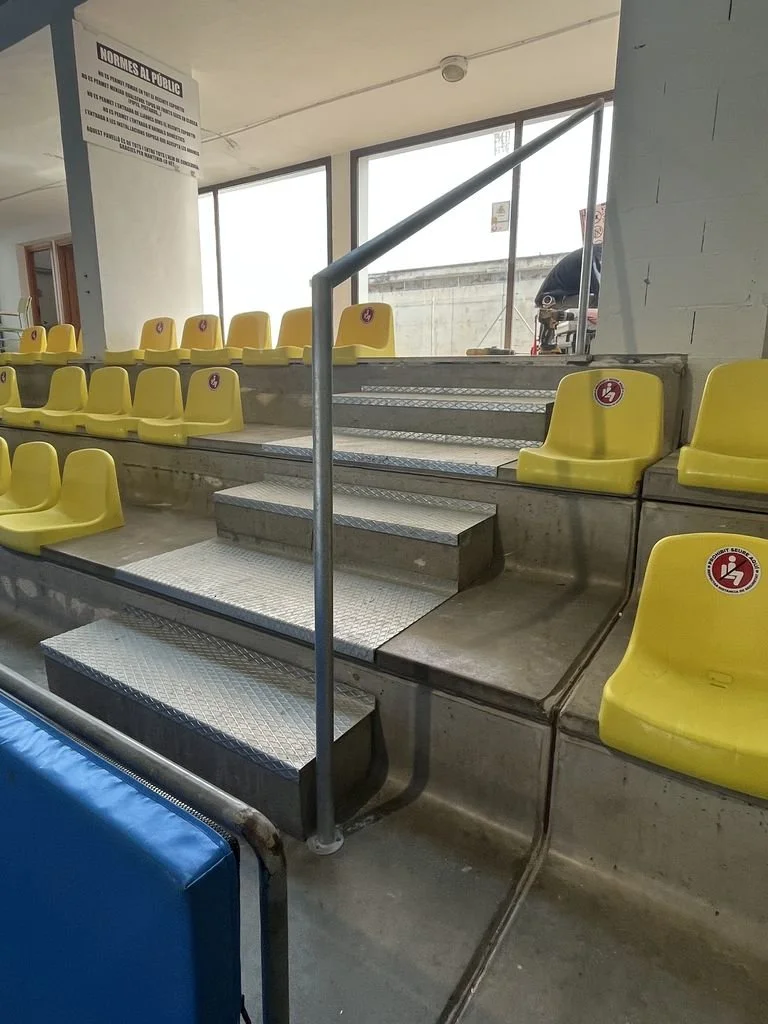 Asientos de estadio amarillos en gradas con escaleras de metal y señalización de distanciamiento social en el área pública.