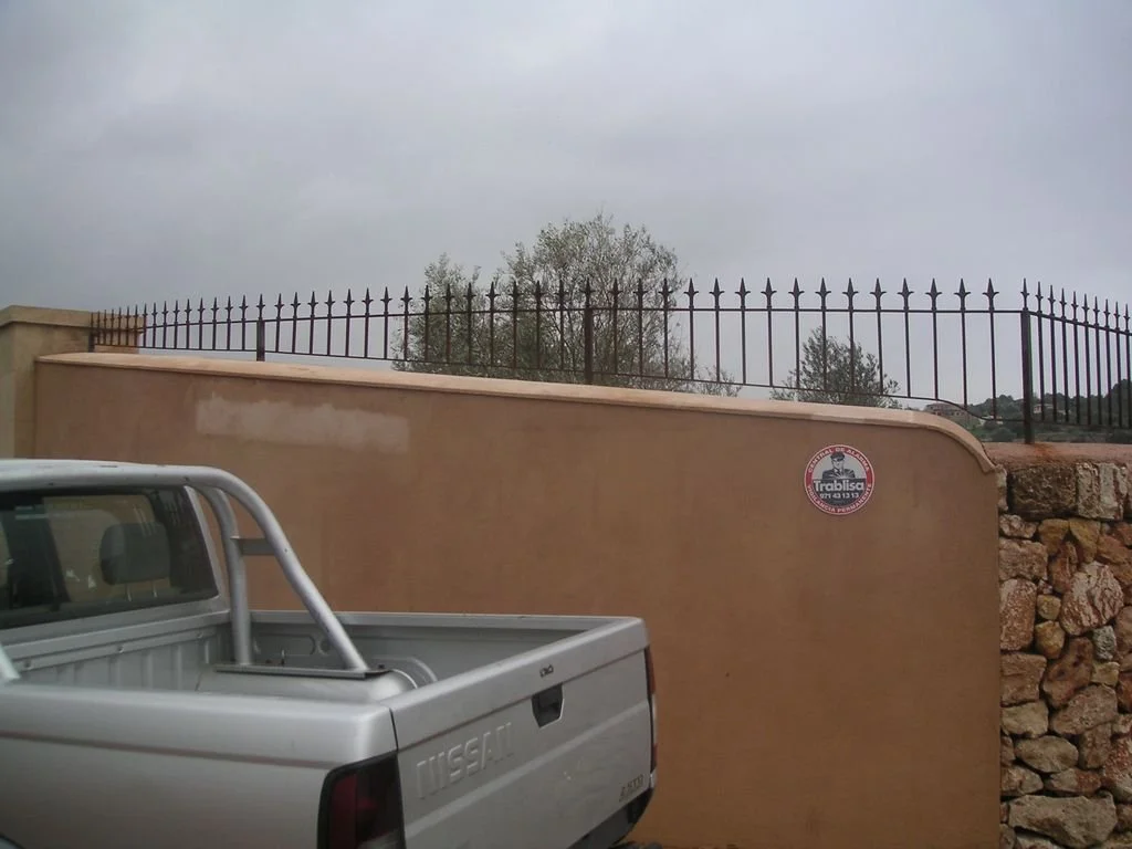 Camioneta gris estacionada frente a una pared de color beige con reja de hierro en la parte superior y una señal de advertencia en la pared, en un día nublado.