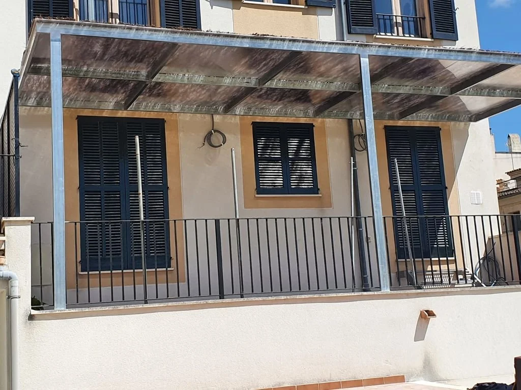 Fachada de una casa con ventanas con persianas negras y un techo de acrílico transparente, con una baranda negra en el balcón.