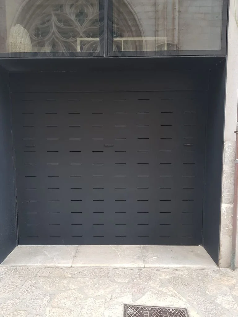 Puerta de garaje negra y moderna con ventanales decorativos arriba, rodeada por paredes de piedra y una superficie de piso de piedra.