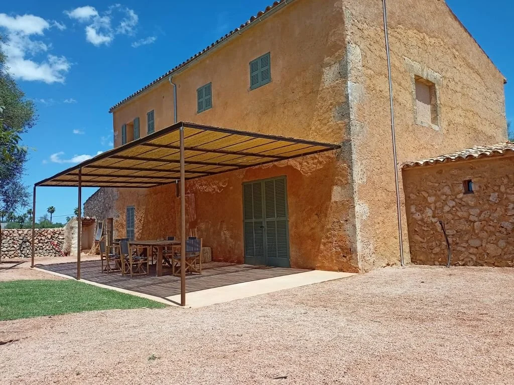 Casa rústica de piedra con patio y estructura de sombra para mesa y sillas en un día soleado.