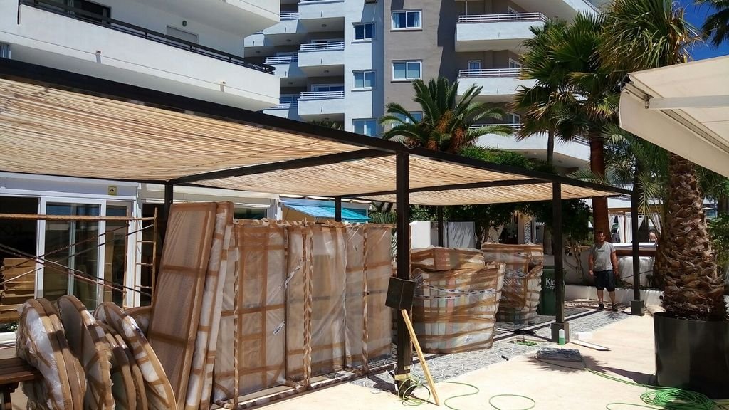 Estructura en una terraza al aire libre, con palmas alrededor, en una zona de construcción con materiales envueltos en plástico y una persona en el fondo.