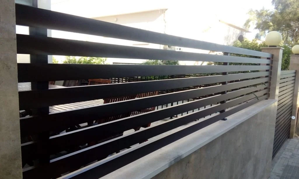 Enrejado negro en una barda de concreto en un área exterior con plantas y luminarias en el fondo.