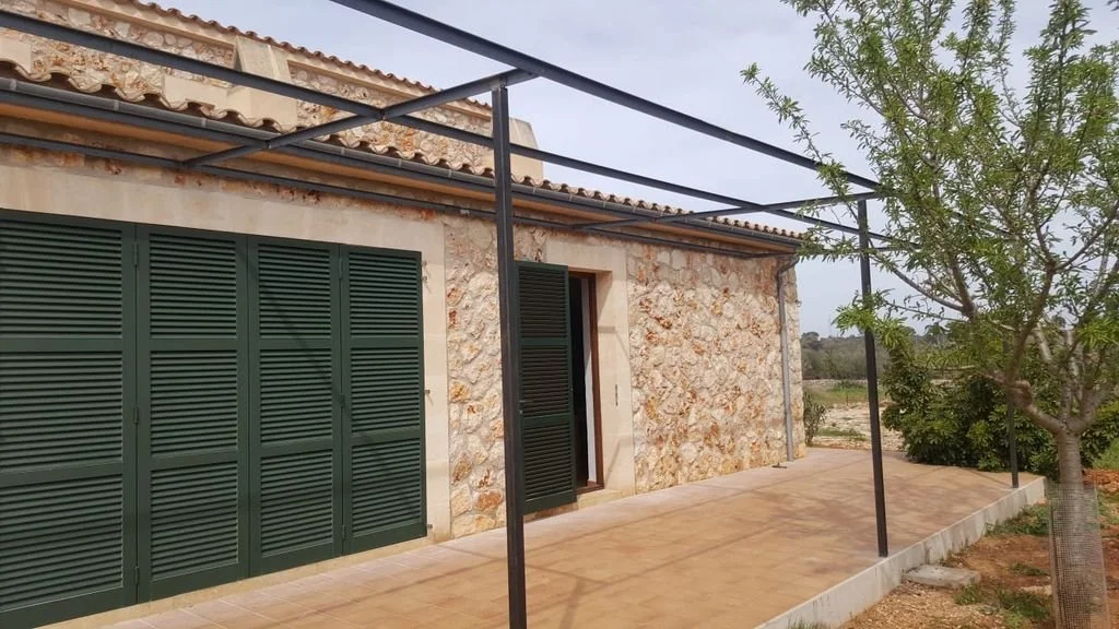 Fachada de una casa con paredes de piedra, ventanas con contraventanas verdes y un patio con piso de baldosas y un árbol a la derecha.