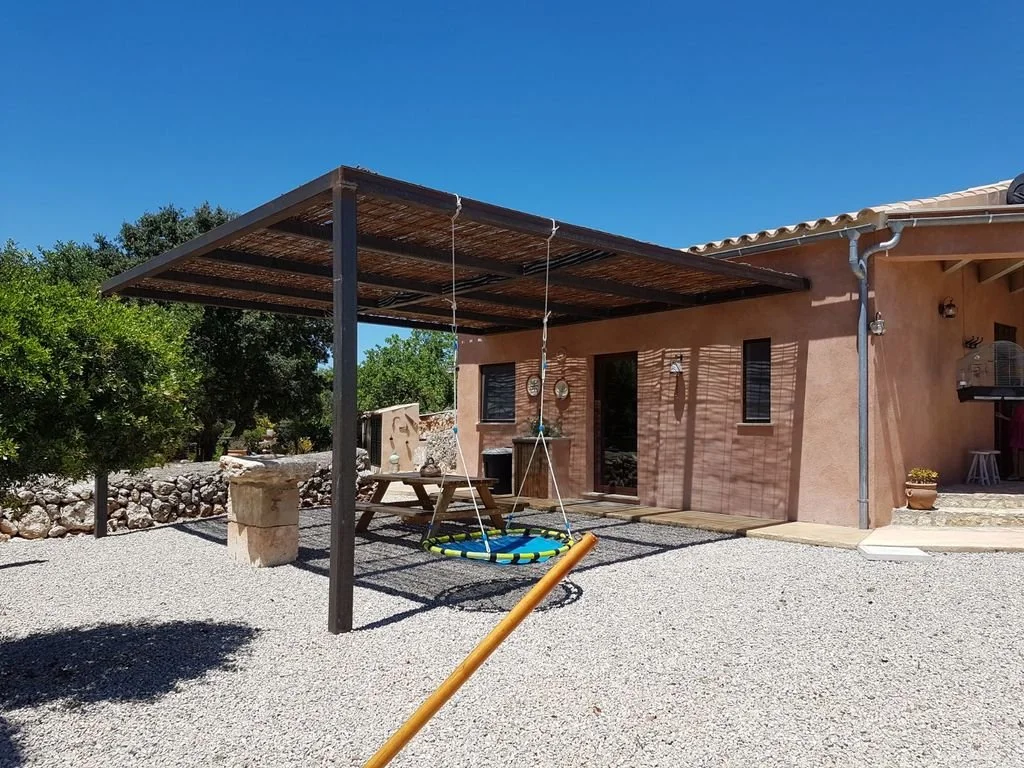 Patio exterior de casa con columpio de ni\u00f1o, mesa de picnic, centro de barbacoa y cobertura de madera bajo cielo despejado