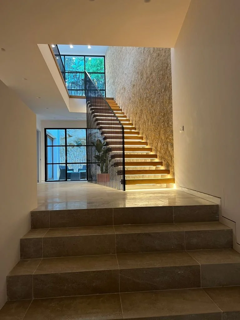 Escalera moderna con iluminación integrada, paredes de piedra y ventanas grandes que permiten la entrada de luz natural.