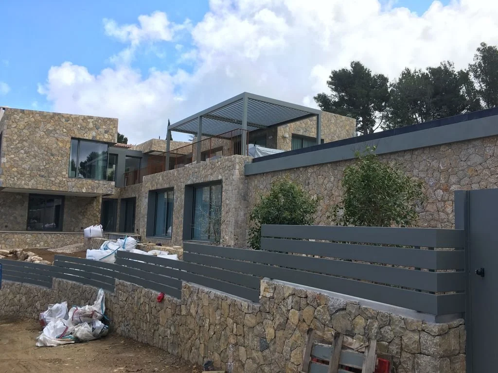 Una casa moderna en construcción con paredes de piedra, ventanas grandes, muro de metal gris y un espacio en la azotea con estructura de techo. Se observan materiales de construcción y árboles en el fondo.
