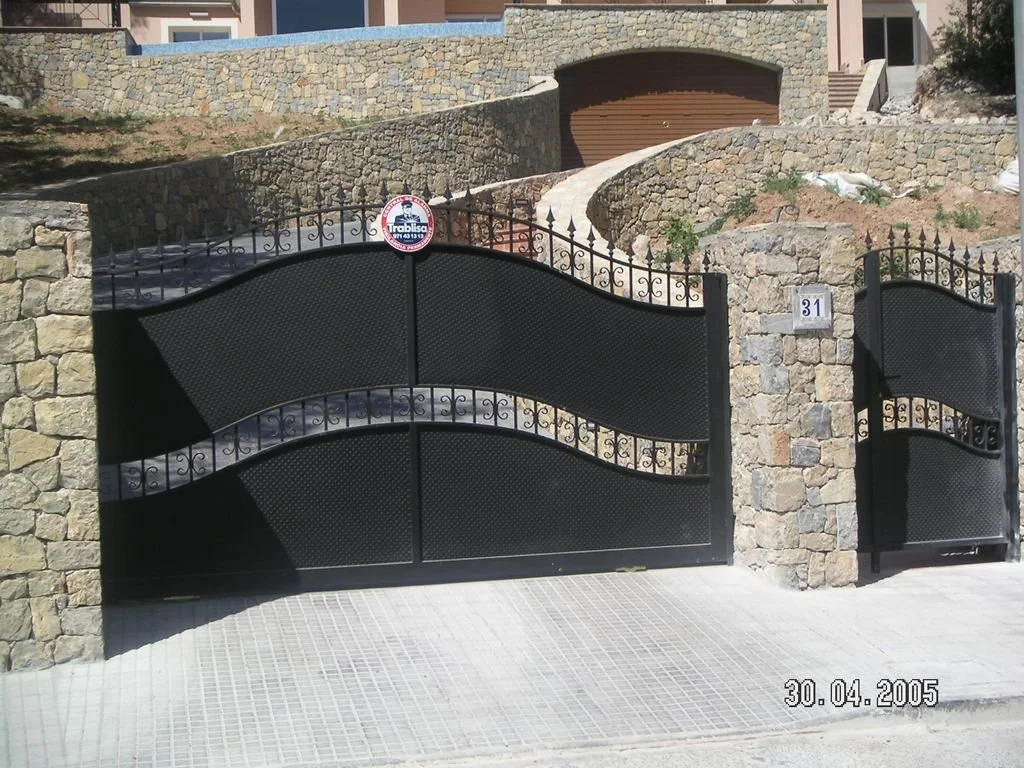 Puerta y portón de hierro negros con muro de piedra y pasillo de piedra en un fondo de vivienda con muros de piedra y escalera.