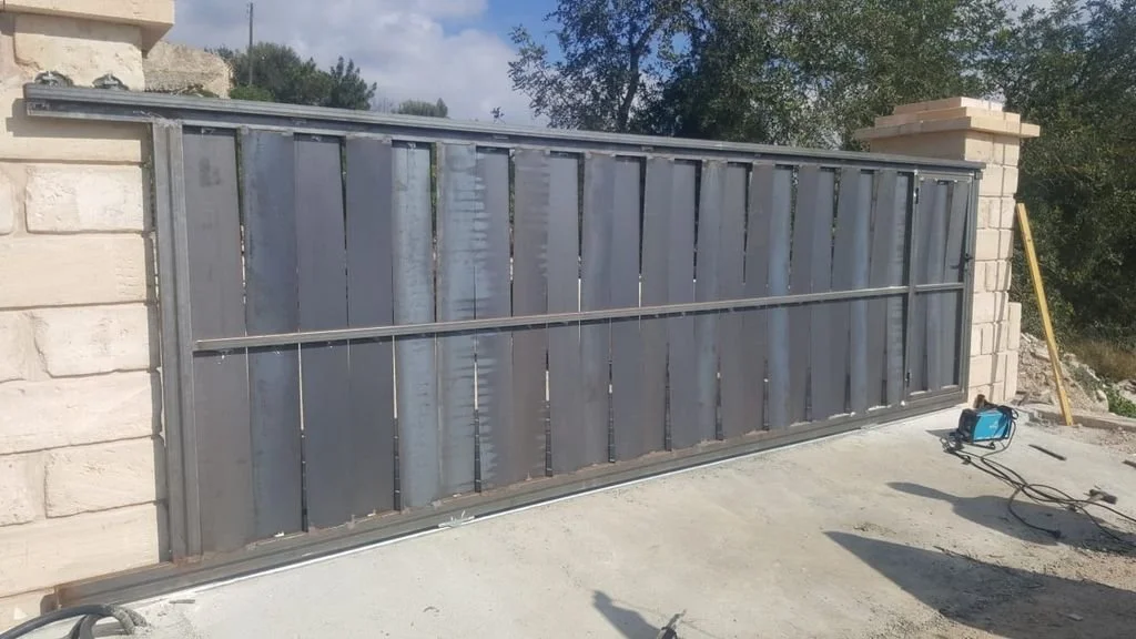 Puerta metálica de cierre en construcción, instalada entre dos columnas de piedra, con herramientas de trabajo a su alrededor, en un entorno exterior.