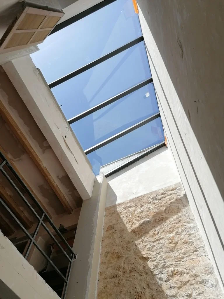 Vista desde abajo de un tragaluz con marco negro y vidrio azul, en un espacio en construcción con paredes de piedra y madera.