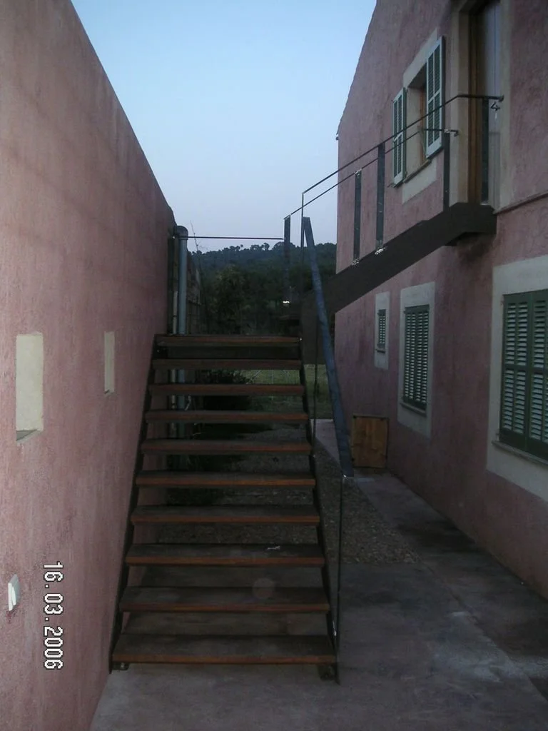 Escalera de madera con pasamanos negro entre dos paredes de color rosa, frente a una casa con ventanas con contraventanas verdes y una pequeña terraza con barandilla.
