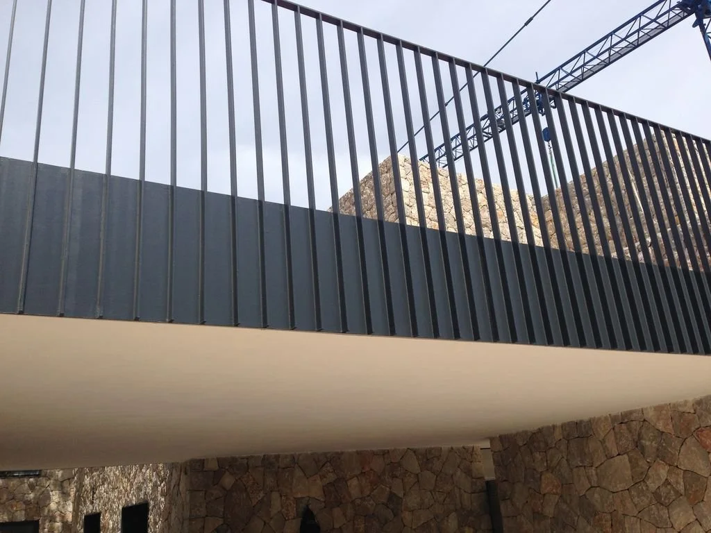 Vista de una estructura moderna con barandal negro, una sección de techo blanco y paredes de piedra, en un entorno montañoso.