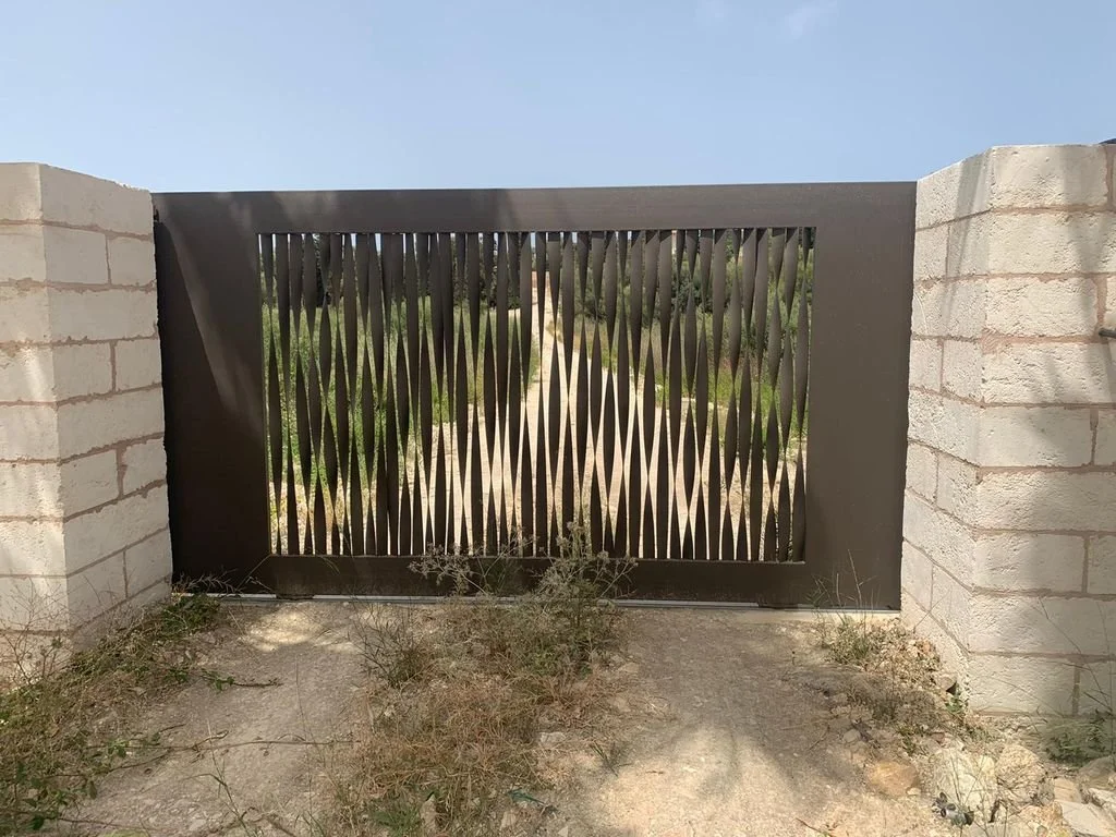 Puerta metálica decorativa con diseño de líneas verticales, entre muros de ladrillo en un entorno exterior con plantas y tierra