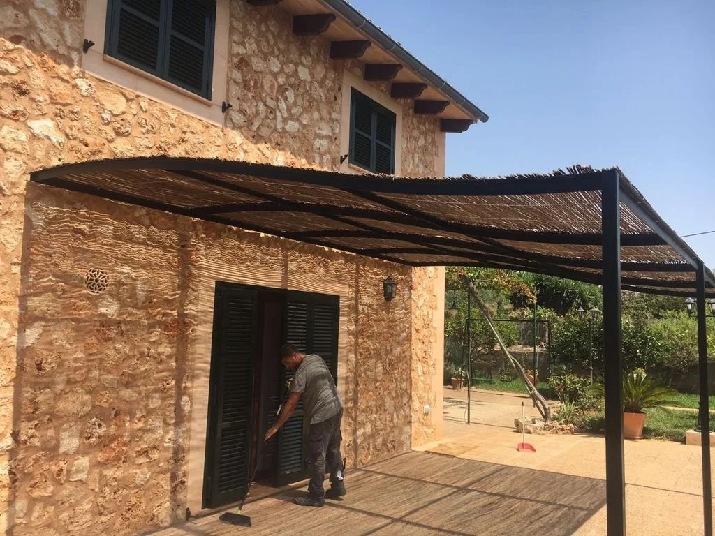 Hombre barriendo la entrada de una casa de piedra con ventanas verdes y un toldo de sombra en el patio.