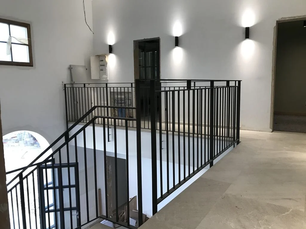Interior de un espacio moderno con barandales negros que rodean una escalera en un segundo nivel, paredes blancas, una ventana y luces de pared.